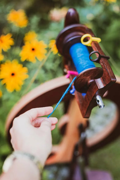 Detailaufnahme der Flyer-Einheit des Fantasia Spinnrads mit blauer Spule vor gelben Blumen im Garten.